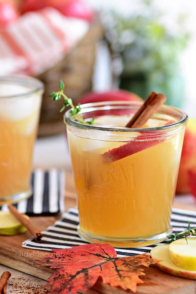 apple cider cocktail with bourbon on a black and white striped napkin and fall leaves and apples around the table