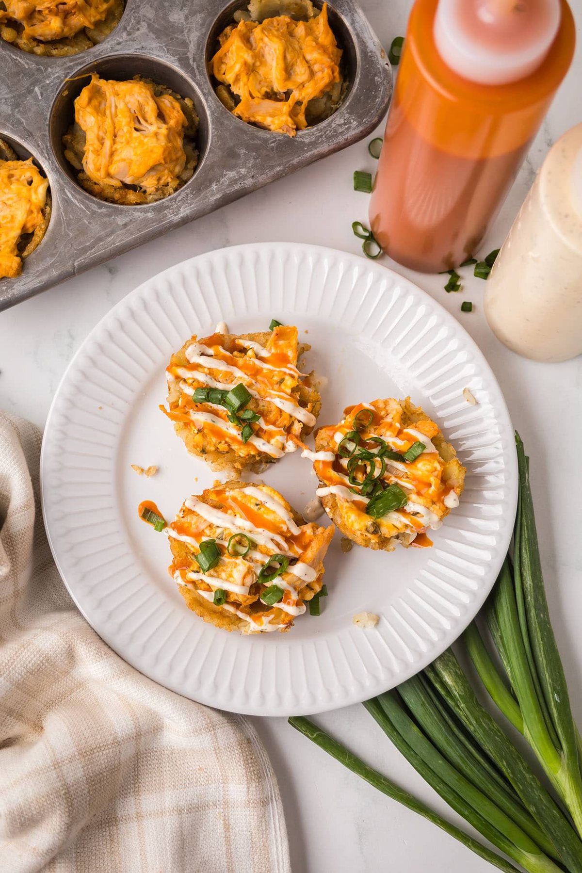 Overhead view of three buffalo chicken tater tot cups topped with ranch drizzle and sliced green onions on a white plate.
