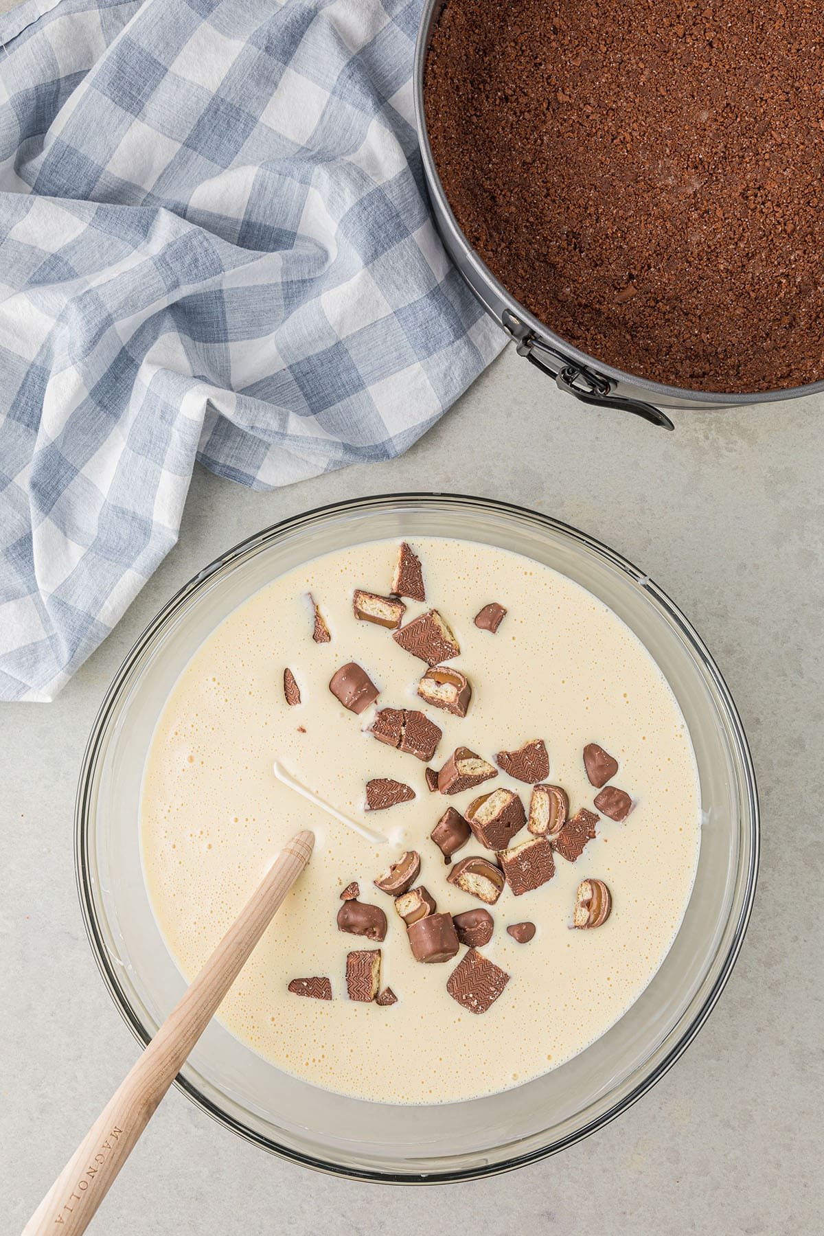 Twix cheesecake batter in a glass mixing bowl and a chocolate cookie crust along side.