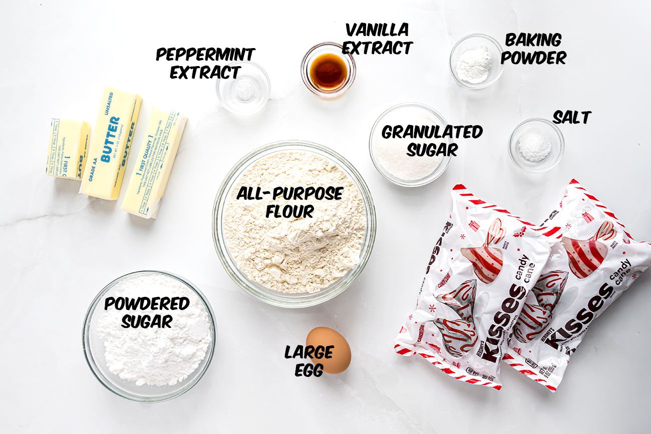 Overhead view of ingredients for Candy Cane Kiss Cookies, including butter, powdered sugar, flour, egg, sugar, peppermint extract, and candy cane Hershey’s Kisses.