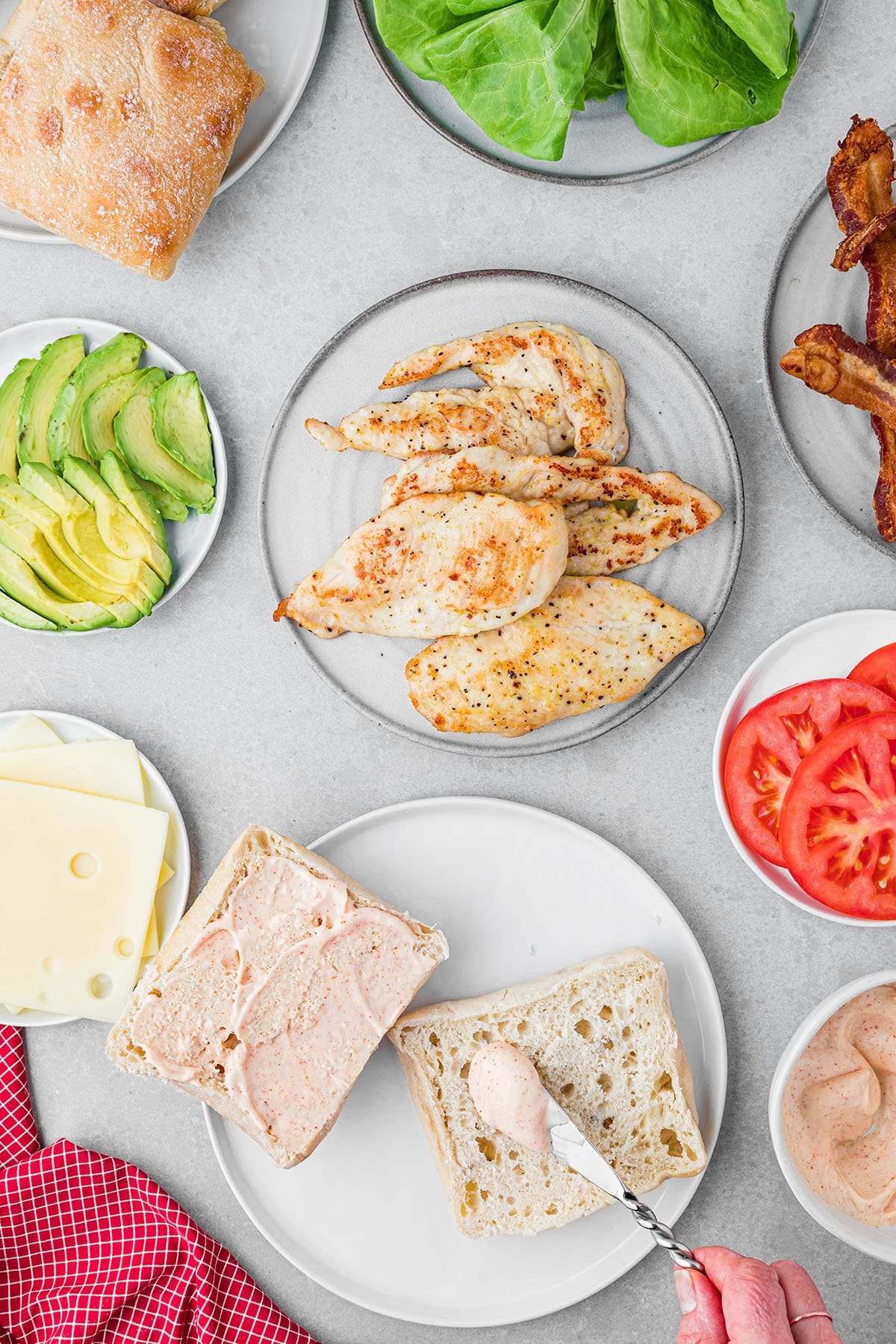 spreading homemade dijonnaise on a sandwich bun with all of the other ingredients plated and around the lunch plate.