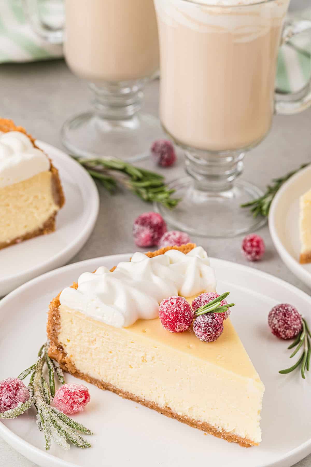 a slice of cheesecake on a white plate with sugared cranberries and a rosemary sprig and 2 cups of eggnog