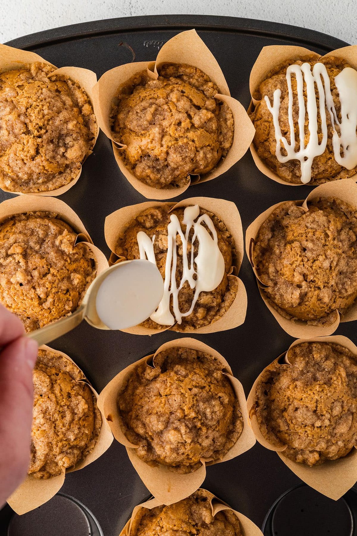 Spoon drizzling maple cream cheese icing over crumble-topped gingerbread muffins in a muffin pan.