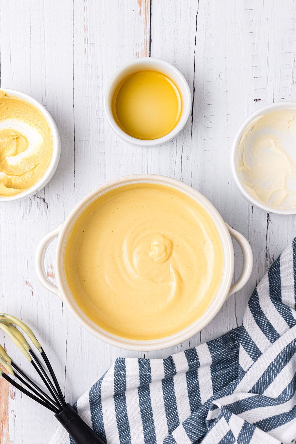 Homemade honey mustard sauce in a small white bowl with a wire wisk and a blue and white towel