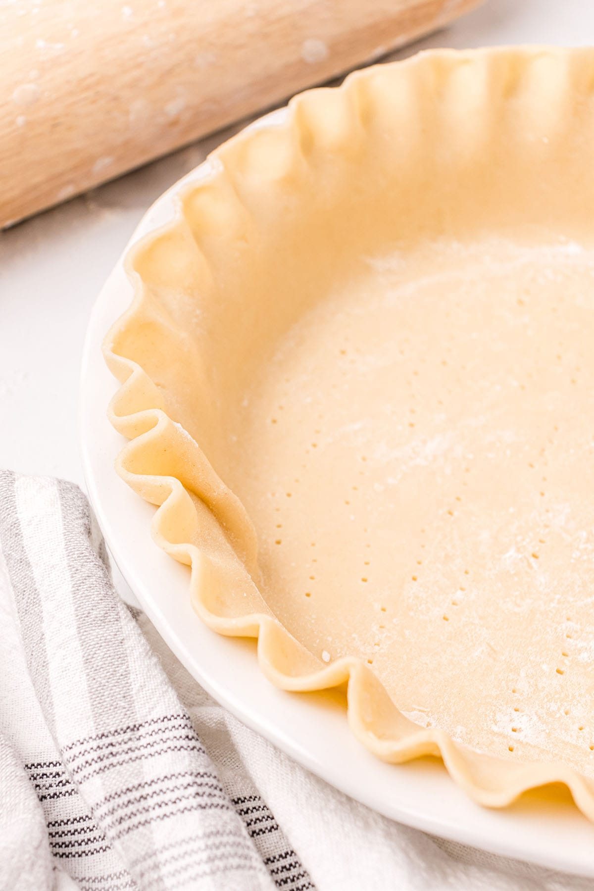 homemade pie crust in a white pie plate with a rolling pin and black and white towel next to it