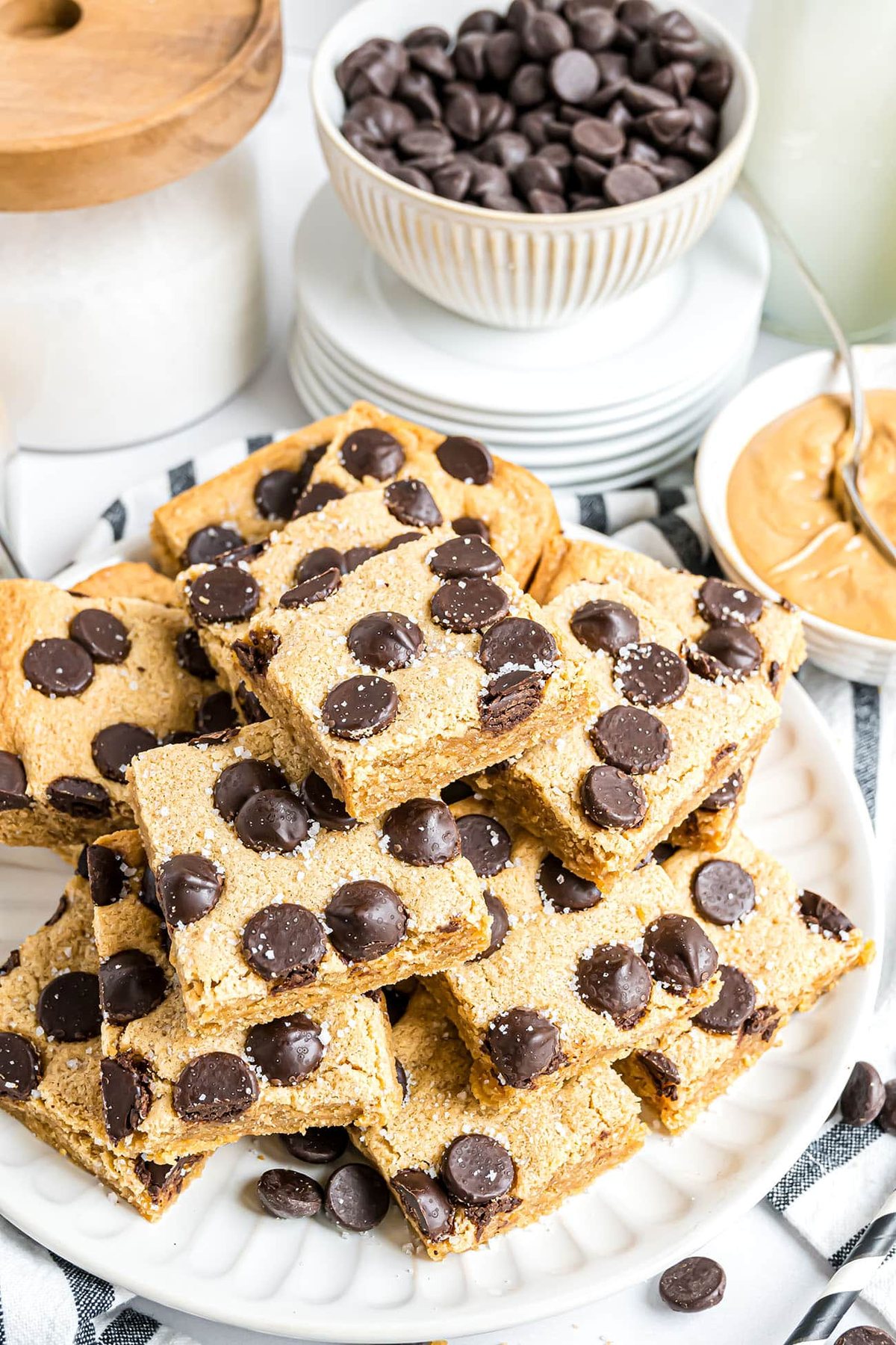 White plate stacked with peanut butter blondies in a pyramid, with chocolate chips and peanut butter bowls blurred in the background.