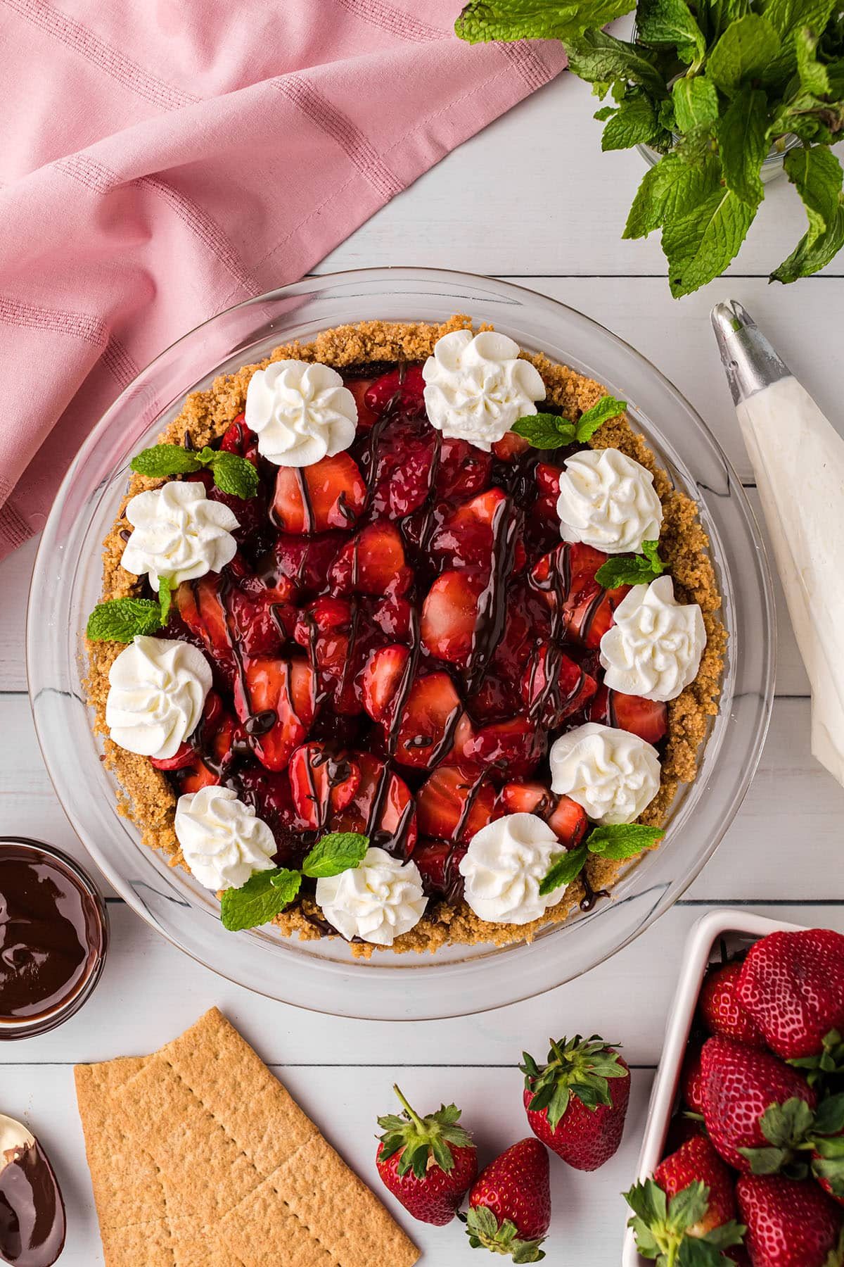 overhead look at No-Bake Fresh Strawberry Cream Cheese Pie with whipped cream and fresh mint garnish on a counter with a pink towel