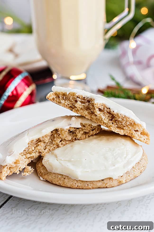 Two perfectly frosted eggnog cookies on a plate, one broken in half revealing its soft, spiced interior.