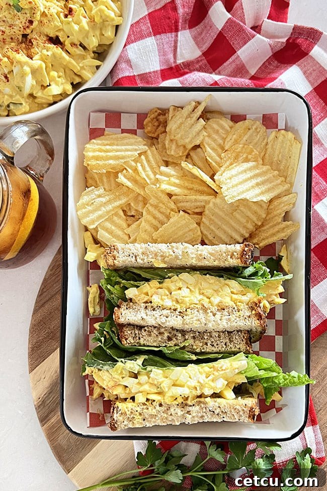 A beautifully presented classic egg salad sandwich in a white tray with crispy potato chips and a red checked napkin.