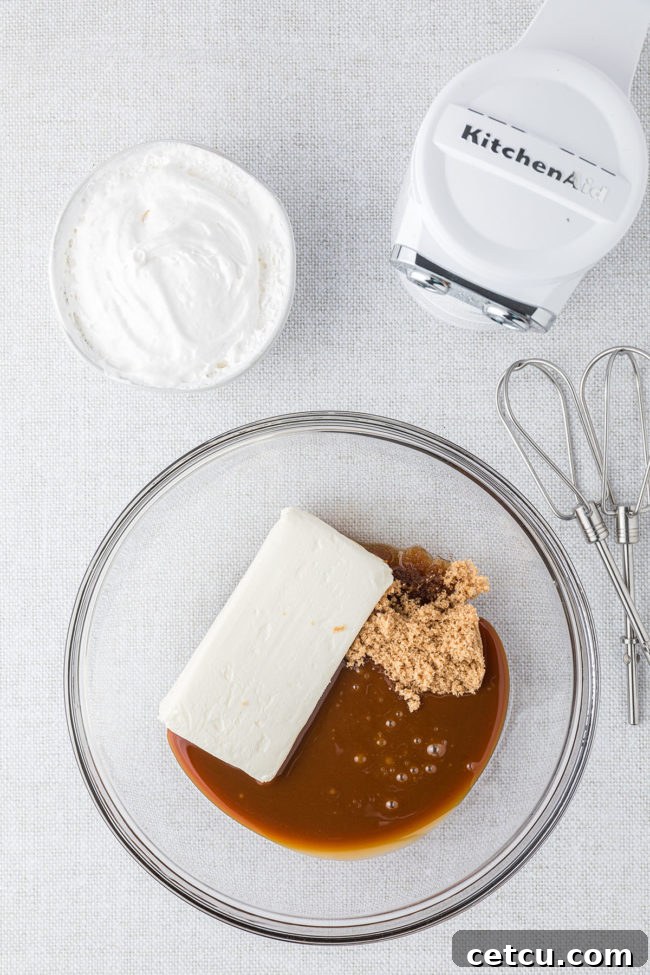 A glass mixing bowl holds softened cream cheese, rich caramel topping, and packed brown sugar, with an electric beater poised to begin mixing.