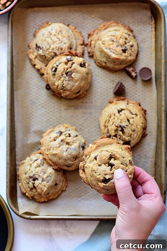 A woman's hand gently holds a freshly baked Peanut Butter Blowout Cookie over a full tray of similar delicious cookies.
