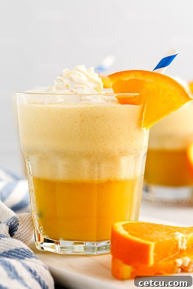 A glass filled with a frothy Homemade Orange Julius, generously topped with whipped cream and a fresh orange slice, sitting elegantly on a kitchen counter.