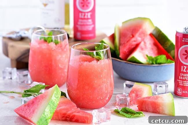 Two stemless wine glasses filled with bright pink Watermelon Grapefruit Wine Slushes, garnished with mint leaves, against a blurred outdoor background. Emphasizes the refreshing nature of the drink.