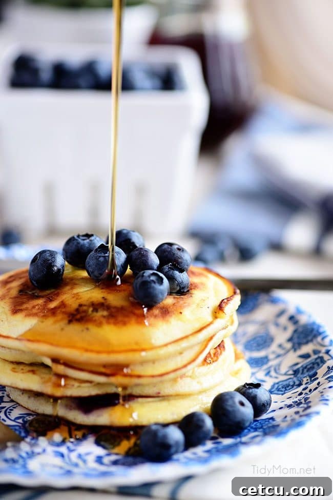 A thick stream of golden maple syrup elegantly cascading over a stack of the Best Homemade Blueberry Pancakes, with vibrant blueberries peeking through, creating an enticing and delicious visual.