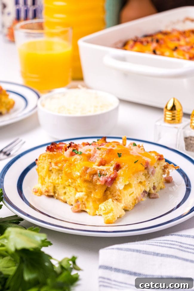 A plate with a serving of English muffin breakfast casserole, accompanied by a casserole dish and a glass of orange juice in the background, set on a wooden table.