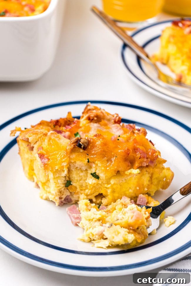 A perfectly portioned serving of English Muffin Breakfast Casserole on a white plate with a fork, showing the fluffy egg, ham, and melted cheese layers.