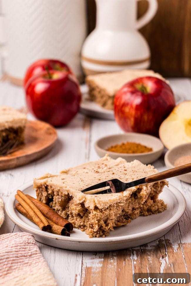 slice of cake on a plate with a fork in the cake and cinnamon sticks. Red apples and cinnamon in the background.