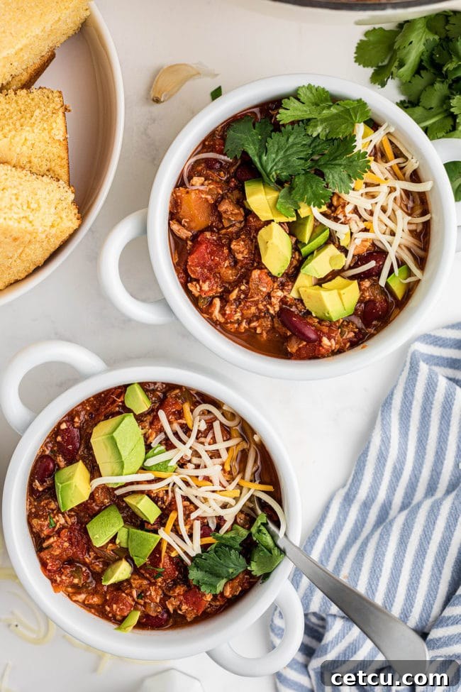 Two bowls of turkey chili artfully topped with creamy avocado slices, shredded cheese, and fresh green herbs.