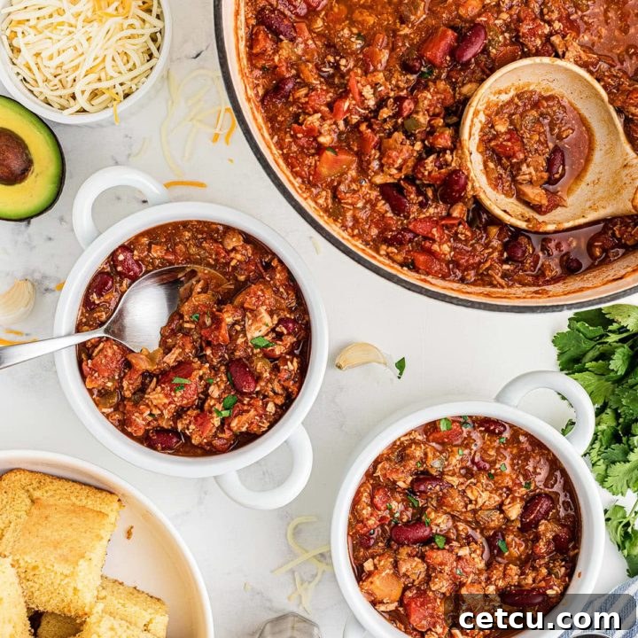 Two white bowls filled with hearty leftover turkey chili, garnished with fresh herbs and a large pot of chili in the background, with cornbread on the side.