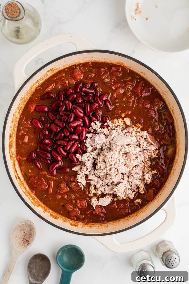 Stirring shredded turkey and dark red kidney beans into the simmering chili base in a large pot.