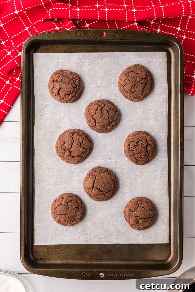 A freshly baked tray of chocolate kiss cookies cooling slightly on a baking sheet, before the Hershey's Kisses are added.