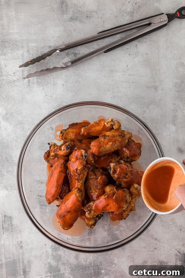 Baked chicken wings being tossed with buffalo sauce in a large glass mixing bowl, ensuring full coating and flavor.