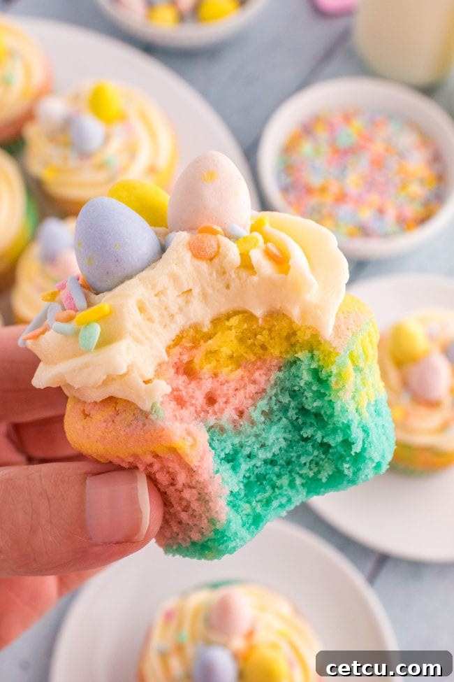 A hand holding a beautifully decorated Spring cupcake with a bite taken, revealing the pastel marble swirl inside, with sprinkles and a mini chocolate egg.