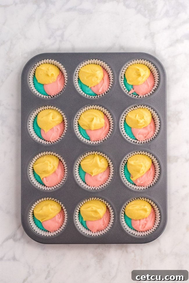 A cupcake pan with liners partially filled with scoops of pastel yellow, pink, and blue cake batter, ready for swirling.