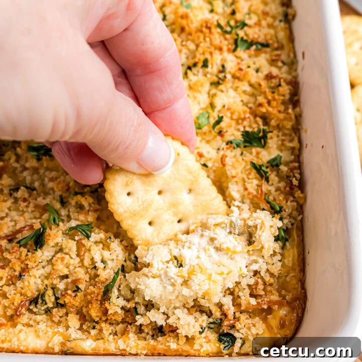 A close-up of a hand scooping warm, creamy jalapeño popper dip with a cracker from a white baking dish, showcasing the golden topping.