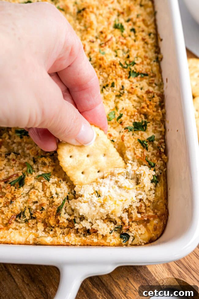 A hand scooping a generous portion of warm, cheesy baked jalapeño popper dip onto a crisp cracker, showing the creamy texture and golden topping.